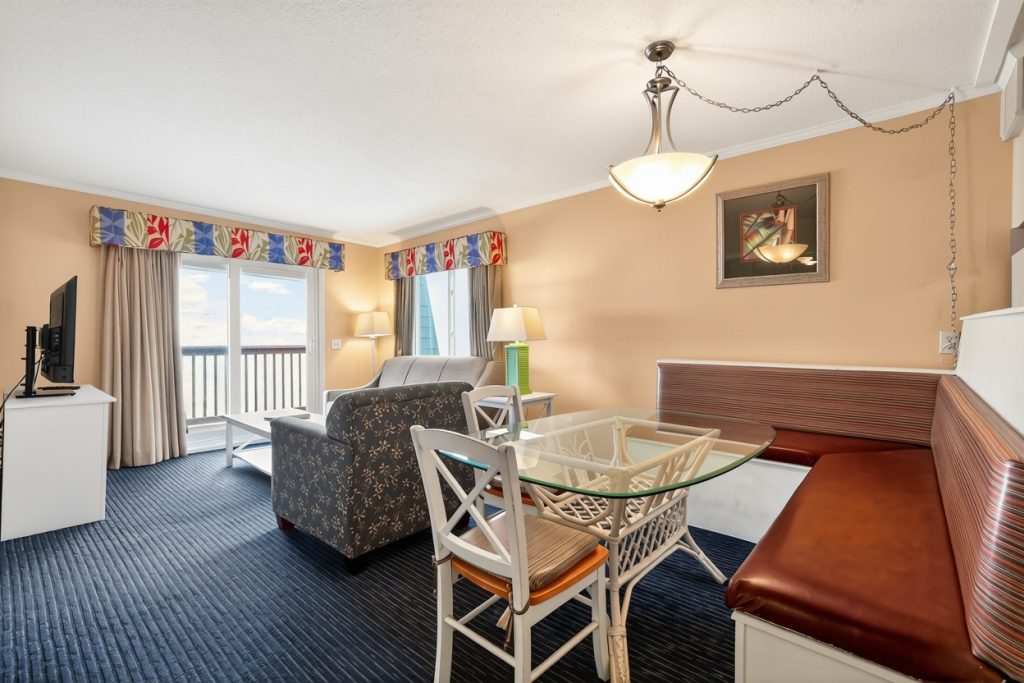 A wide view of a hotel room with a sofa, dining table, TV, and balcony.